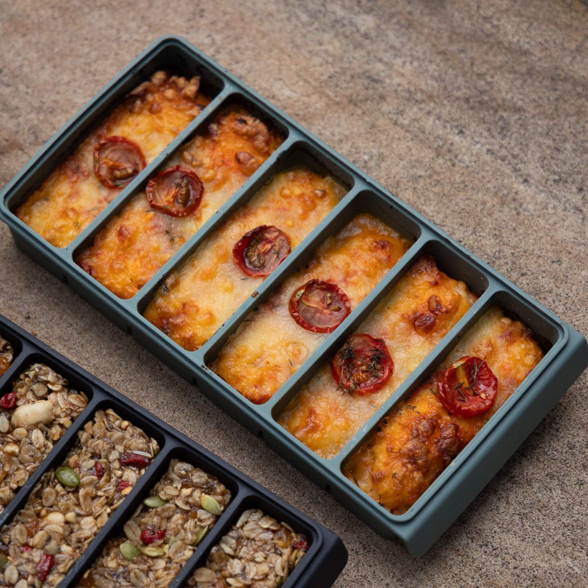 Close-up of a eucalyptus green 6-portion tray filled with baked cheesy tomato portions, with a second tray of oat-based bars partially visible — showing oven-to-table use