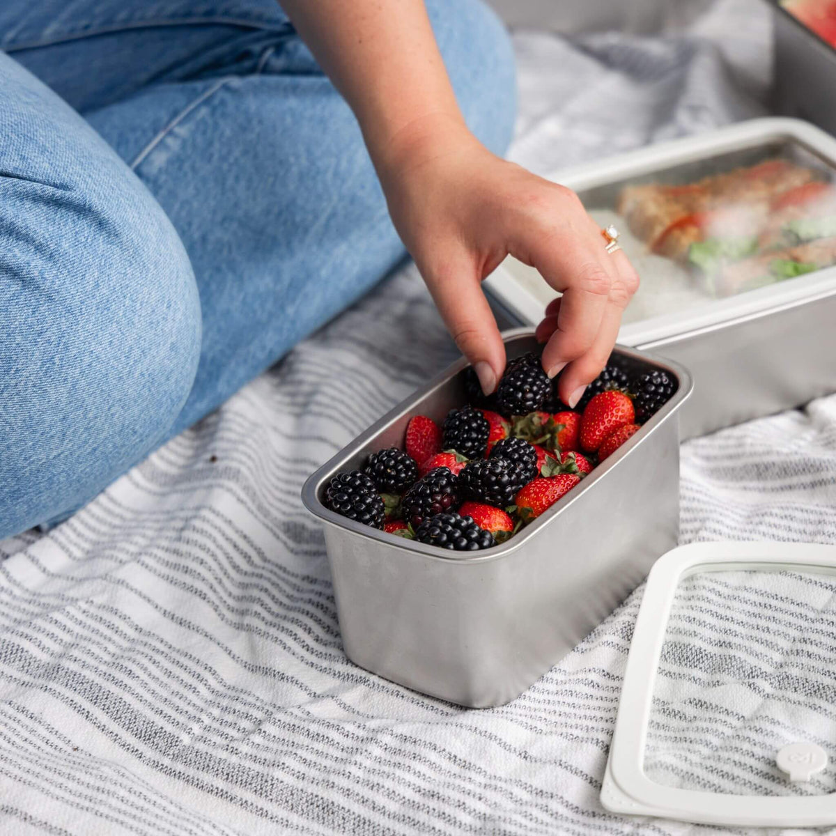 Hand reaching into an open Rectangle Stainless Steel Container filled with strawberries and blackberries at a picnic, with the glass lid set aside and a second container with food visible in the background.