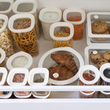 A pantry drawer with different glass food containers that have white lids. The containers contain breads, grains, snacks and condiments